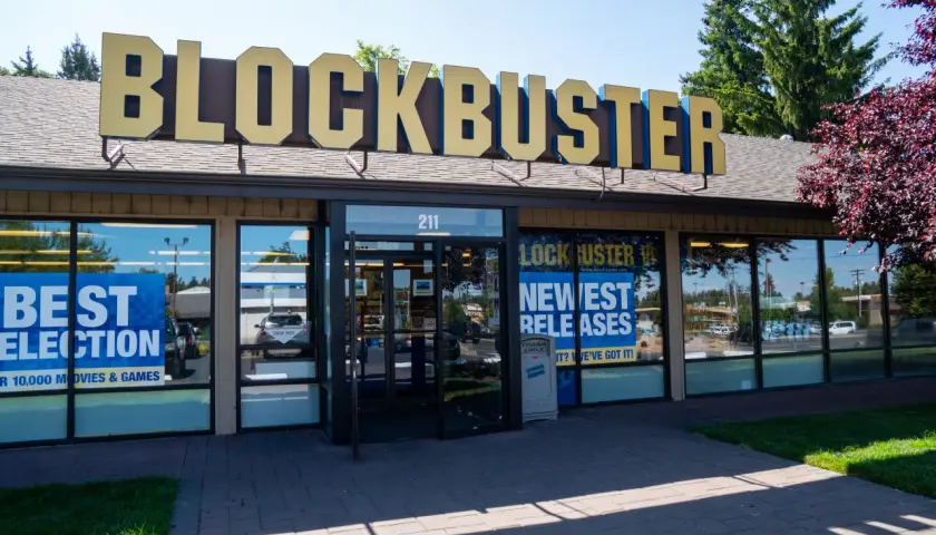 L'insegna dorata del negozio Blockbuster domina l'edificio. Vetrine mostrano "Best Selection" e "Newest Releases", un richiamo nostalgico ai tempi d'oro del blockbuster film.