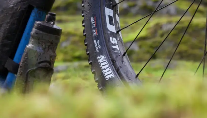 Close-up of a muddy bike tire, showing the "Maxxis Ikon" branding. This tire is ready for a challenging trail review.