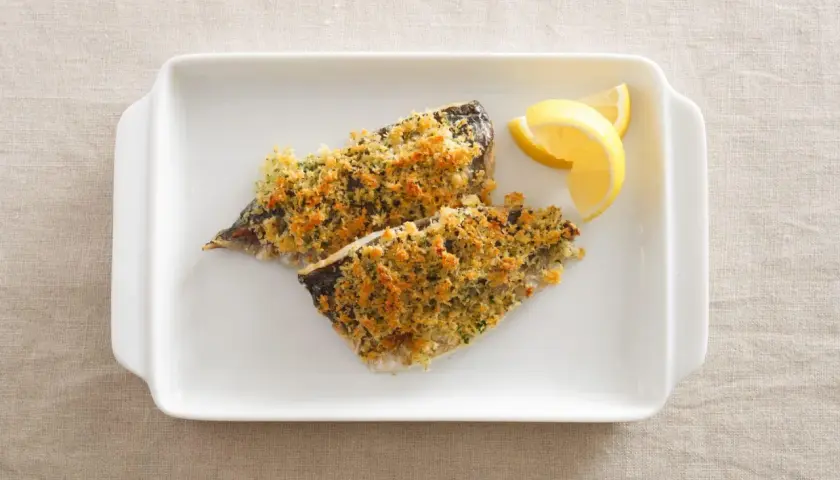 Two fillets of aji fish, baked with a golden breadcrumb topping, served with lemon wedges on a white dish.