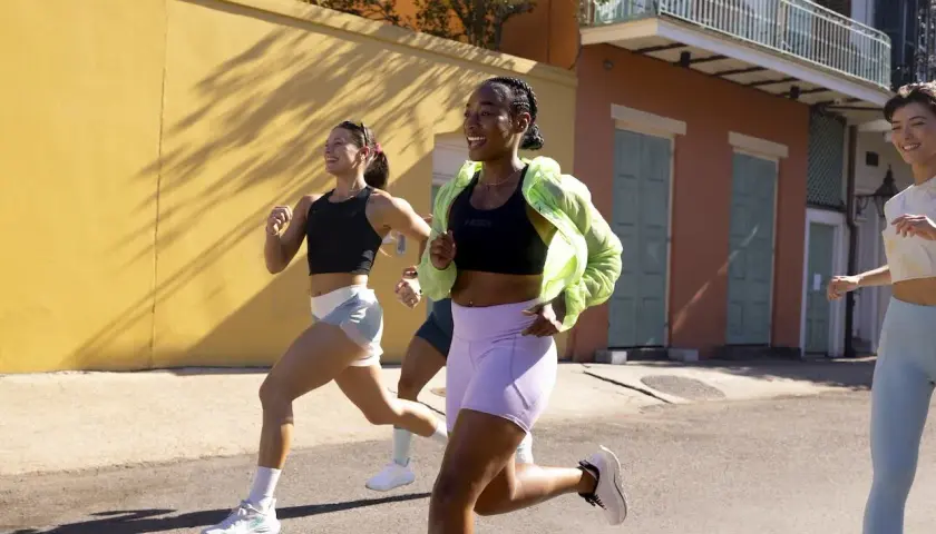 Trzy kobiety w sportowych strojach biegną ulicą. Jedna z nich, z nadwagą, uśmiecha się radośnie, pokonując kolejne metry.