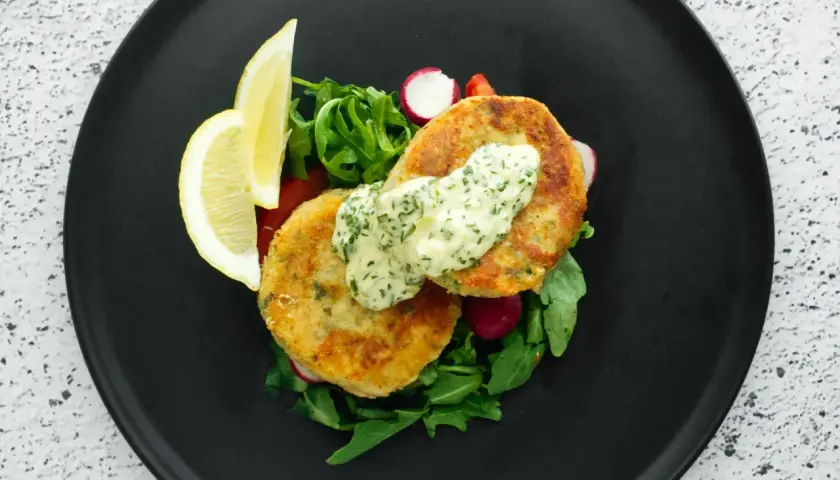 Golden chikuwa tempura patties served with a creamy herb sauce, fresh greens, radishes, and lemon wedges on a black plate.