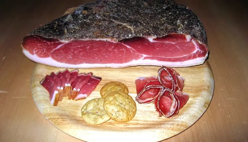Ein Stück italienischer Speck mit Scheiben und Keksen auf einem Holzbrett.