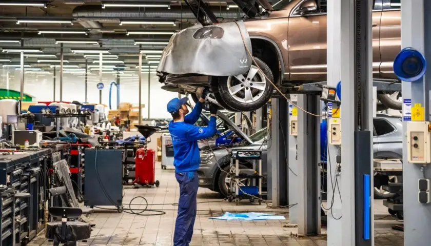 Mekaniker arbetar under en bil på en lyft i en auktoriserad verkstad. Verkstaden är välutrustad med verktyg och maskiner.