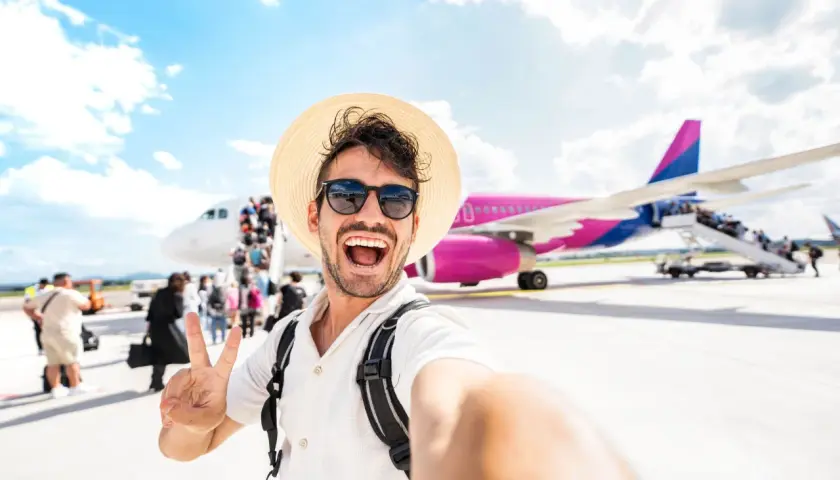 Mężczyzna w słomkowym kapeluszu i okularach przeciwsłonecznych robi selfie na tle samolotu i pasażerów.