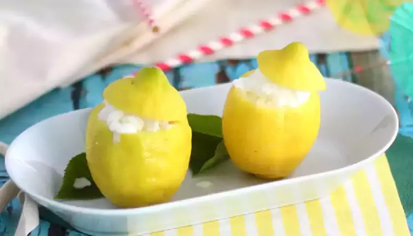 Deux citrons évidés remplis de sorbet citron, servis sur une assiette blanche, avec des feuilles vertes et des pailles colorées en arrière-plan.