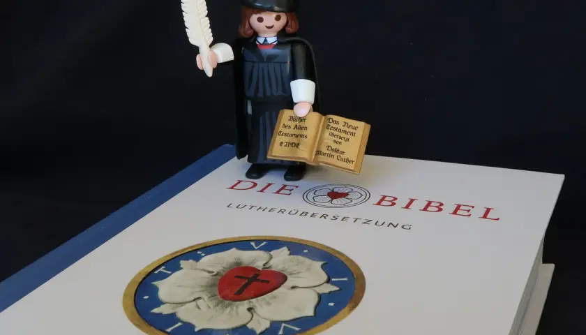 Playmobil-Figur mit Federkiel und Büchern, die den Unterschied zwischen Lutherbibel und katholischer Bibel symbolisiert.