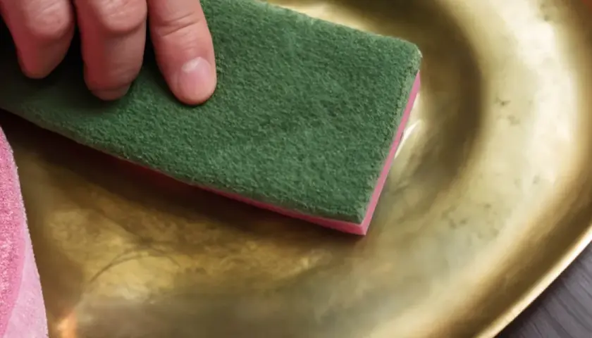 Preparazione per verniciare infissi alluminio dorato: una mano lucida una superficie dorata con una spugna abrasiva verde.