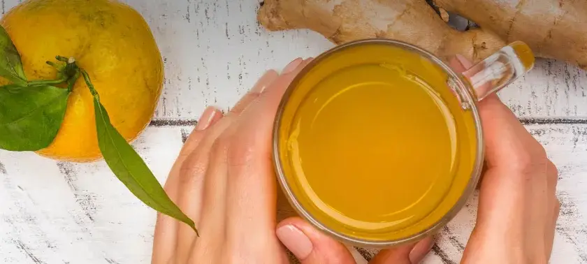 Hände halten ein Glas mit leuchtend gelbem Getränk. Daneben liegen eine Orange und Ingwer. Perfekt, um Kurkuma zuzubereiten.