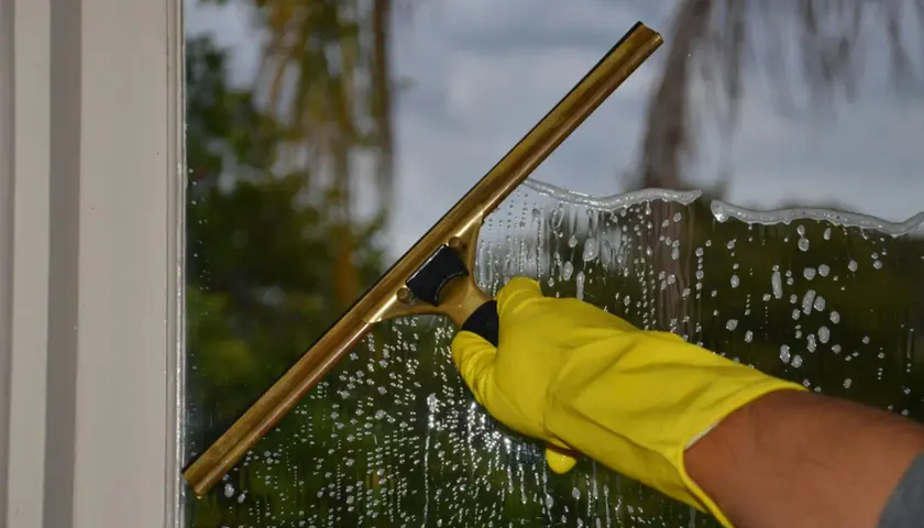 Mano con guanto giallo usa un tergivetro per pulire i vetri delle finestre, lasciando una scia di schiuma e gocce.