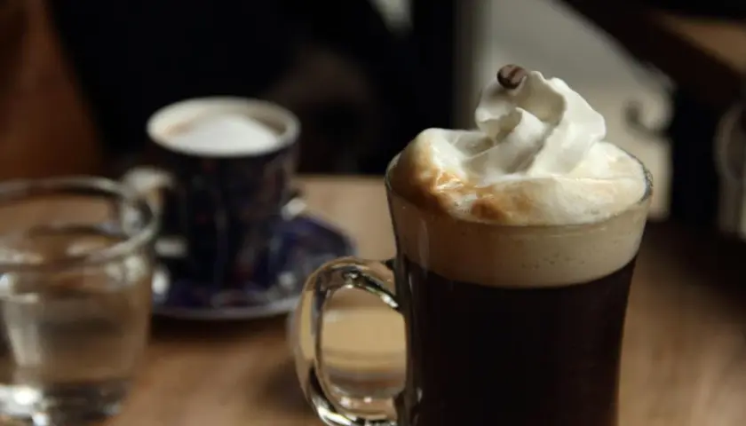 Ein Glas mit Kaffee und Sahne, garniert mit einer Kaffeebohne. Daneben ein kleiner Becher und ein Glas Wasser. Perfekt für einen Kaffee mit Schnaps.