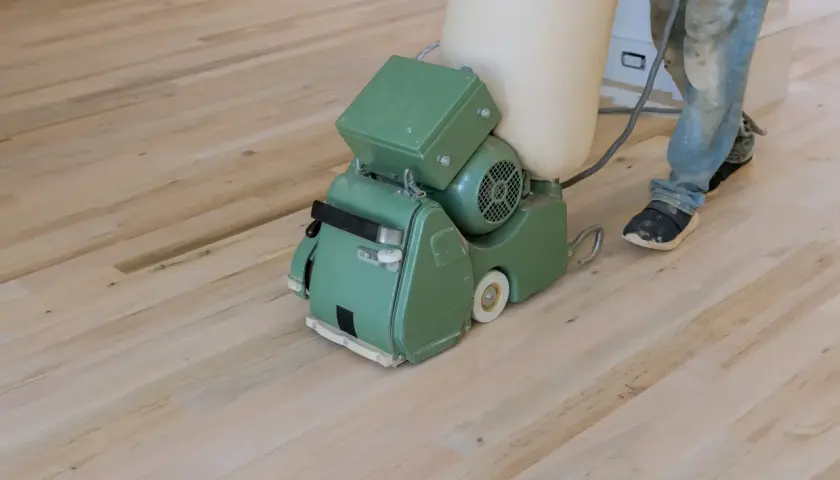 Ein Handwerker schleift mit einer Maschine einen hellen Dielenboden. Staub wird in einem Beutel gesammelt.