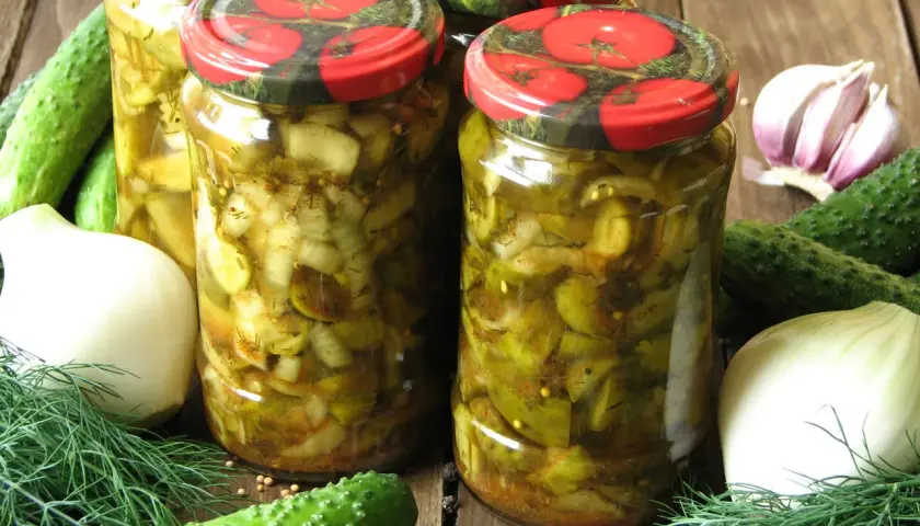 Słoiki z ogórkami w curry ze skórką, otoczone świeżymi ogórkami, cebulą, czosnkiem i koprem.