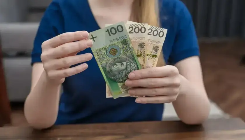 Kobieta trzyma banknot 100 zł i dwa po 200 zł. Łącznie 500 zł, czyli połowa kwoty 1000 plus.