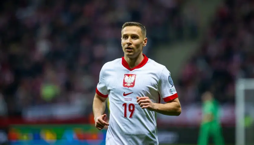 Frankowski piłkarz w białej koszulce z orłem na piersi, numer 19. Biegnie po boisku, skupiony na grze.