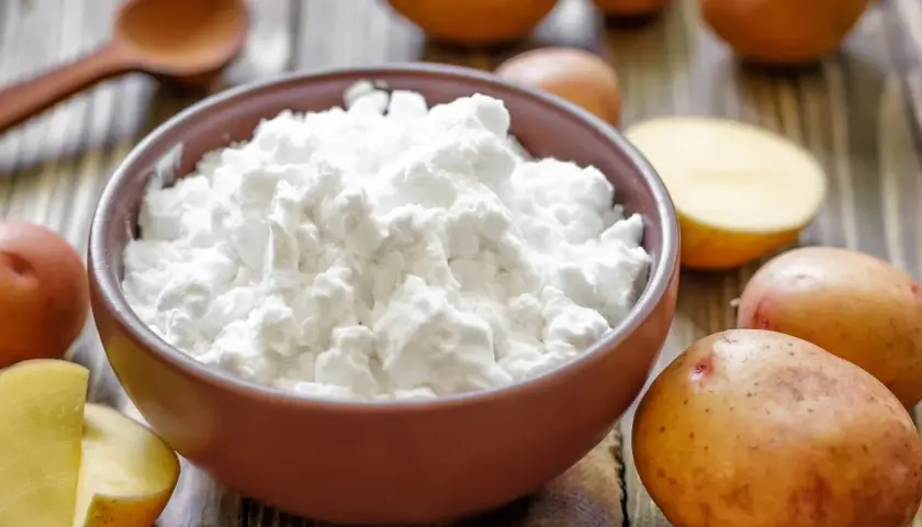 Miska pełna białego krochmalu ziemniaczanego, obok ziemniaki i drewniana łyżka. Idealne składniki do domowej kąpieli w krochmalu.