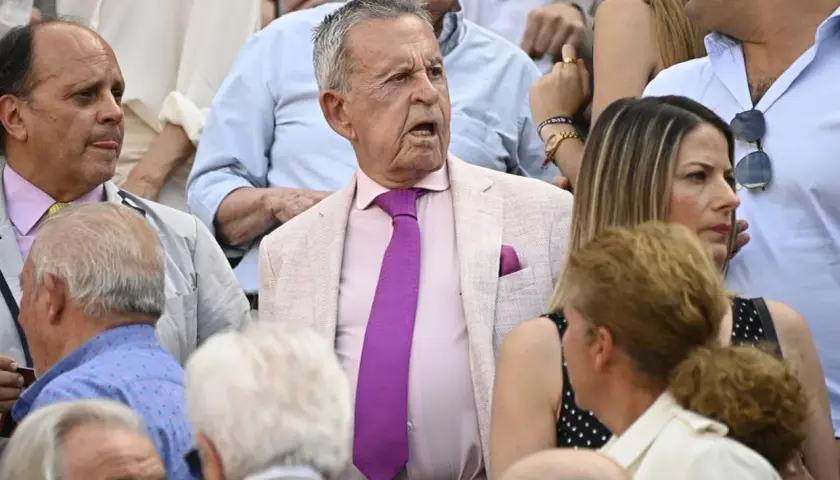 Un grupo de personas, entre ellos algunos famosos en las ventas, observan atentamente un evento. El hombre del centro, con traje rosa y corbata morada, parece sorprendido.