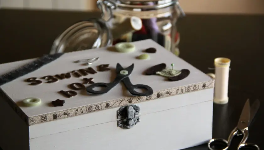 Un costurero pequeño decorado con letras, tijeras de fieltro y botones. Un tarro lleno de hilos y tijeras completan la escena.