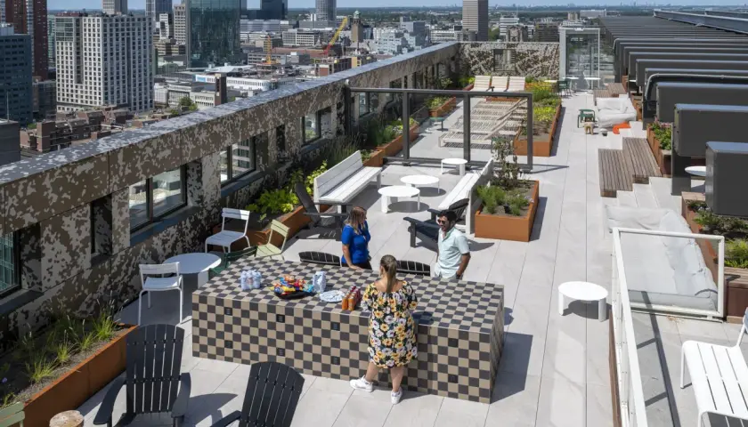 Ein begehbarer Flachdachgarten mit Stadtblick. Menschen entspannen sich auf der Dachterrasse, die mit Sitzgelegenheiten, Pflanzen und einem Schachbrett-Tisch ausgestattet ist.