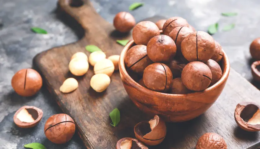 Noci macadamia in una ciotola di legno, con alcune sparse sul tagliere. Scopri i noci macadamia benefici per la tua salute.