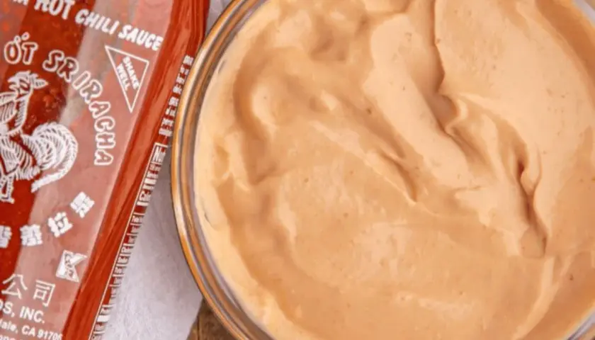 A bowl of creamy, orange spicy mayo recipe sits next to a bottle of Sriracha hot chili sauce.
