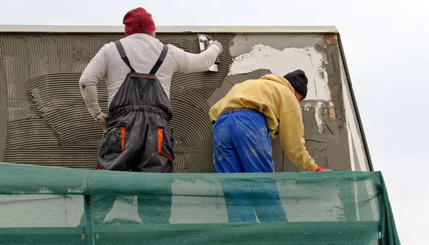 Lavoratori applicano rasatura armata con rete in fibra di vetro su un muro esterno, protetti da un telo verde.