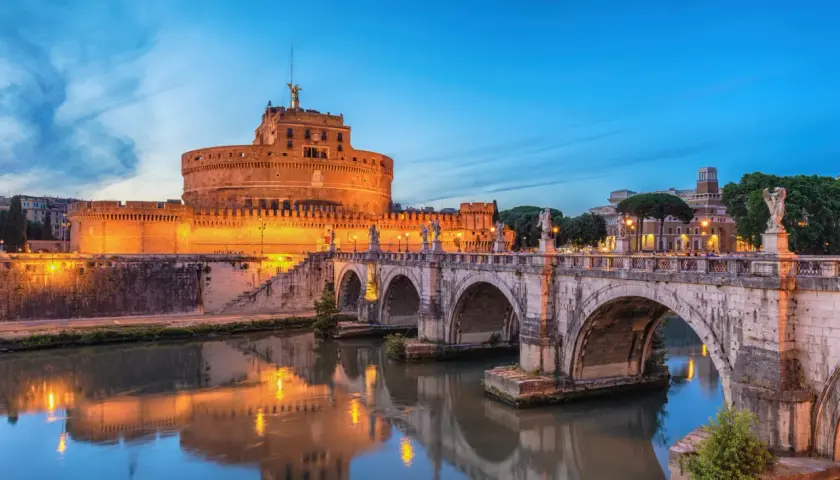Le Castel Sant'Angelo à Rome, illuminé au crépuscule, se reflète dans le Tibre. Le pont Saint-Ange, orné de statues, mène à cette ancienne forteresse.
