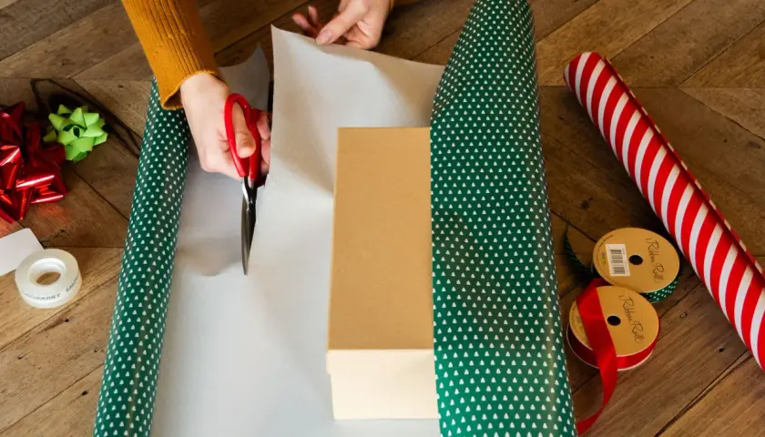 Hände schneiden Geschenkpapier zu, um ein Geburtstagsgeschenk zu verpacken. Rote Schere, Geschenkband und Schleifen liegen bereit.