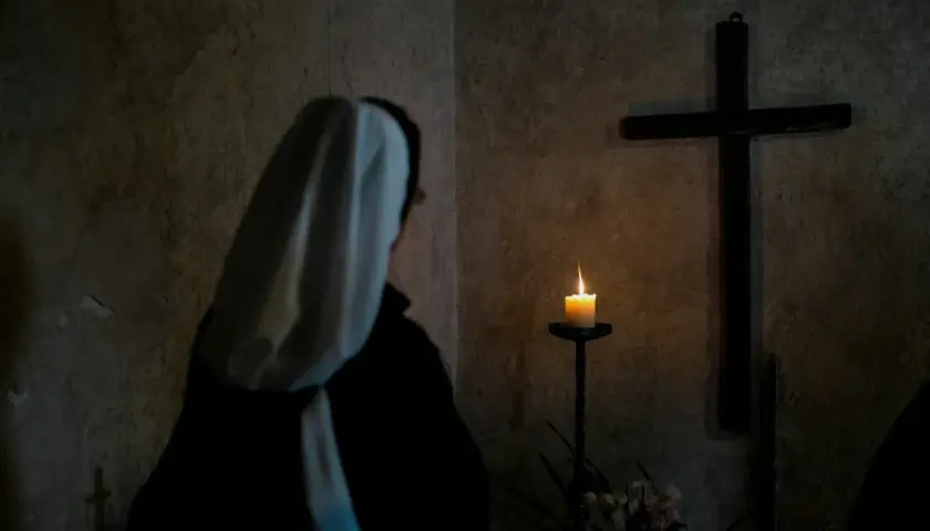 Zakonnica w białym welonie patrzy na płonącą świecę i krzyż w ciemnym pomieszczeniu.