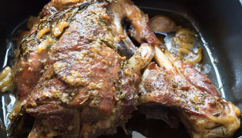 Une succulente épaule d'agneau confite, dorée et parfumée, baignant dans son jus avec des herbes et de l'ail.