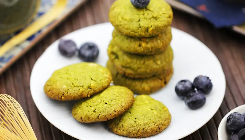 Grüne matcha kekse mit Blaubeeren auf einem Teller, daneben ein Matcha-Schneebesen.