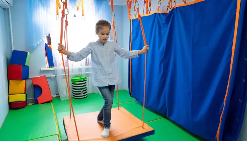 Dziewczynka na huśtawce w sali do terapii sensorycznej. Terapia sensoryczna na czym polega? Na stymulacji zmysłów poprzez ruch i zabawę.