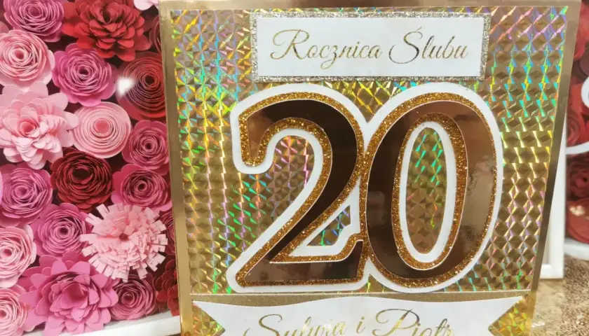 Złota kartka z napisem "Rocznica Ślubu" i liczbą "20" dla Sylwii i Piotra. Obok ramka z różowymi papierowymi kwiatami.
