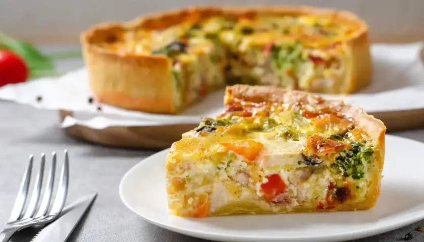 Une part de quiche savoureuse avec une pâte à tarte maison dorée, garnie de légumes et de poulet. Le reste de la tarte est flou en arrière-plan.
