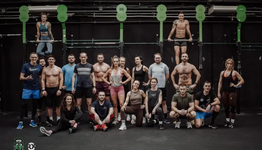 Grupa osób w strojach sportowych pozuje przed czarną ścianą w siłowni CrossFit.