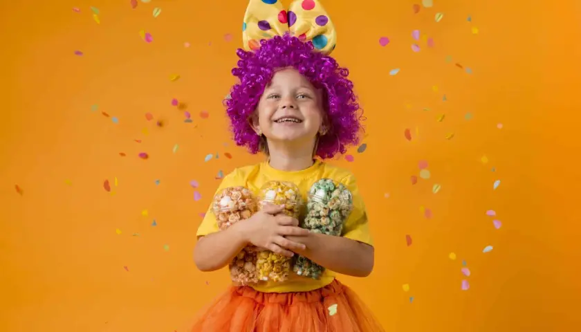 Niña con disfraz casero de payaso, peluca morada, lazo amarillo con lunares y falda naranja, sosteniendo tres vasos de palomitas de colores.