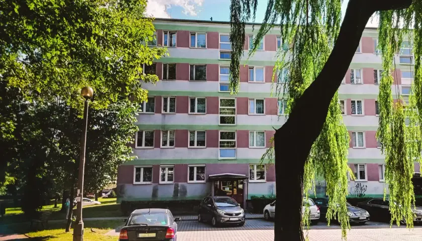 Blok mieszkalny z zielonymi i różowymi pasami, drzewa i zaparkowane samochody.