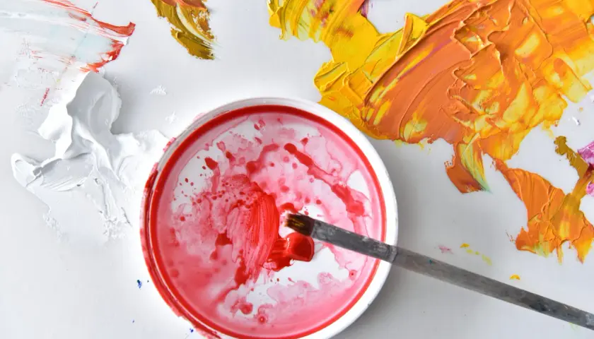 Ein Pinsel taucht in rote Farbe. Daneben sind gelbe und orangefarbene Farbkleckse. So mischt man rot.