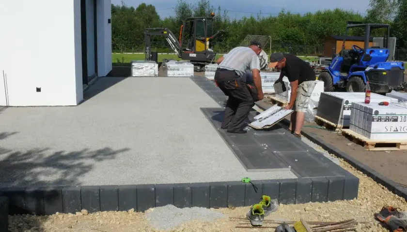 Pracownicy układają płyty na tarasie betonowym. W tle koparka i maszyna budowlana.