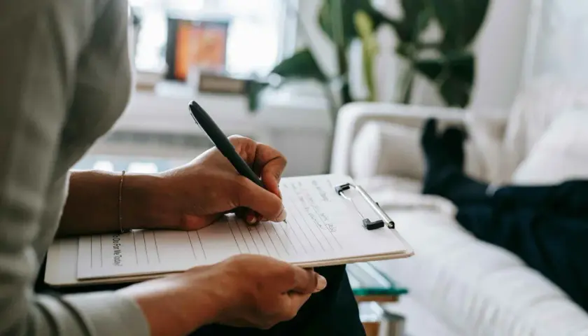 Mani che scrivono su un blocco note, mentre una persona è sdraiata su un divano. Un momento di riflessione per capire se per l'ansia meglio psicologo o psichiatra.