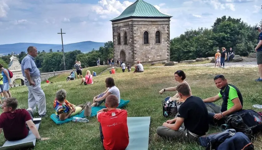 Ludzie odpoczywają na trawie pod kamienną wieżą i krzyżem. Trasa na Święty Krzyż przyciąga turystów.