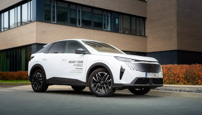 Biały samochód na e, nowy Peugeot 3008 Hybrid, zaparkowany przed nowoczesnym budynkiem z ceglaną elewacją.