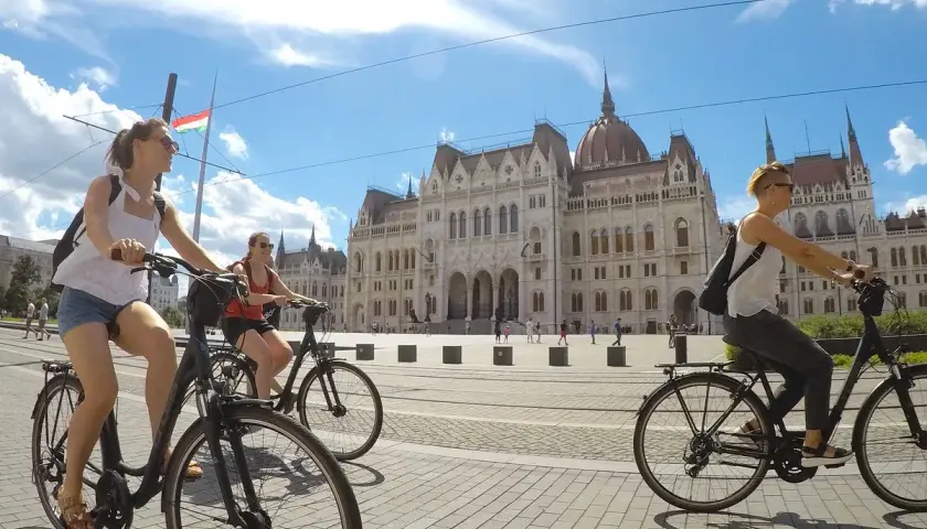Friends enjoying Budapest bike tours, cycling past the Parliament building under a bright, cloudy sky.
