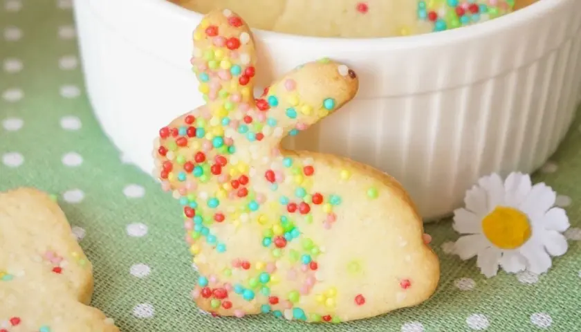 Bunte Osterkekse zum Ausstechen, verziert mit bunten Streuseln, liegen auf einem grünen Tupfentuch. Ein süßer Osterhase steht im Vordergrund.