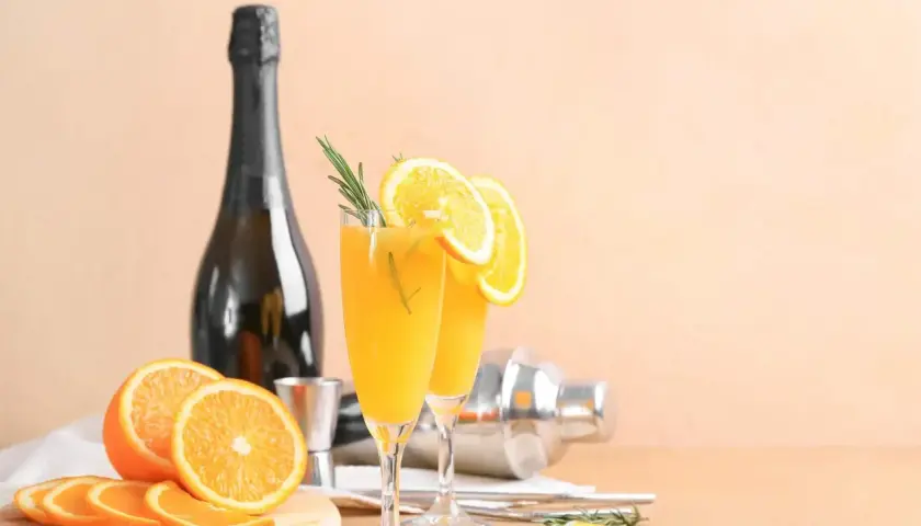 Dwa kieliszki z pomarańczowym drinkiem, ozdobione plasterkami pomarańczy i rozmarynem. Obok butelka, shaker i świeże pomarańcze. Wódka łza pszenicy idealnie pasuje do takiego orzeźwienia.