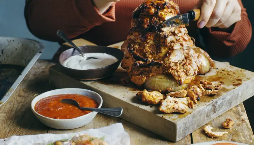 En person skär av en bit från en saftig kyckling shawarma som roterar på ett spett. Tillbehör som vitlökssås och en röd sås finns bredvid.