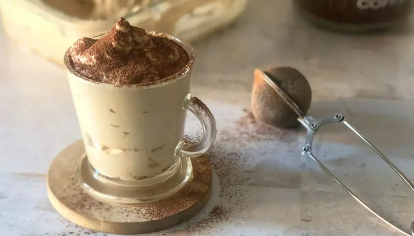 Deliziosa crema al caffè in una tazza di vetro, spolverata di cacao. Accanto, un colino per dolci e un barattolo di caffè.