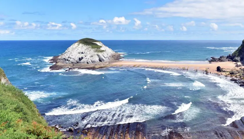 Olas rompen en las **playas limpias de Cantabria**, con un islote rocoso y vegetación verde.