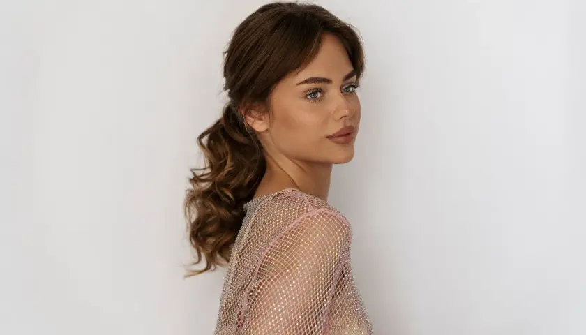 Semirecogidos peinados de madrina media melena: una mujer con cabello castaño ondulado recogido en una cola baja, luciendo un vestido brillante.