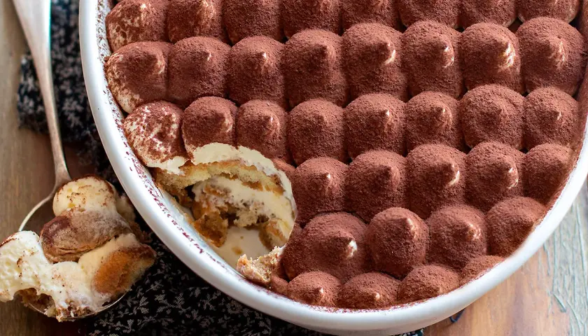 Un invitante tiramisù in una teglia bianca, cosparso di cacao. Una porzione è già stata servita su un cucchiaio, mostrando gli strati cremosi.