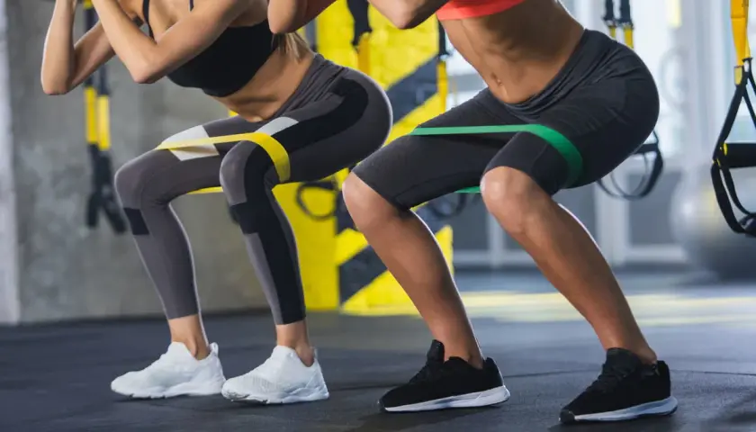 Dwie kobiety wykonują przysiady z gumami oporowymi na udach. Ćwiczenie to angażuje mięśnie podobnie jak maszyna hack squat, wzmacniając nogi i pośladki.